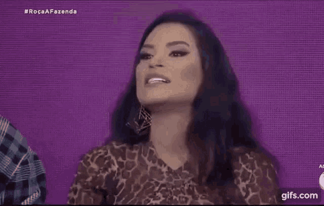 a woman in a leopard print dress is sitting in front of a purple background and waving her hands .