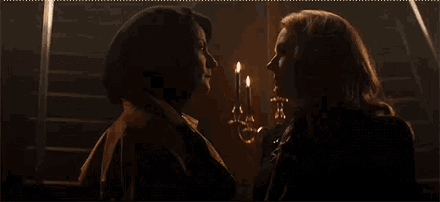two women are looking at each other in a dark room with candles in the background .