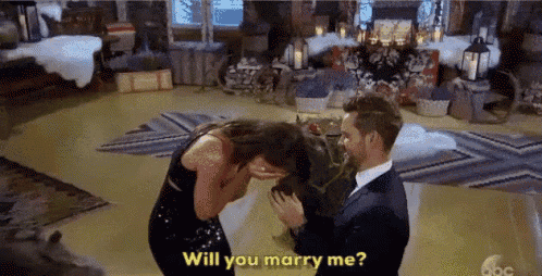 a man is proposing to a woman in a room and the woman is crying and asking him to marry her .