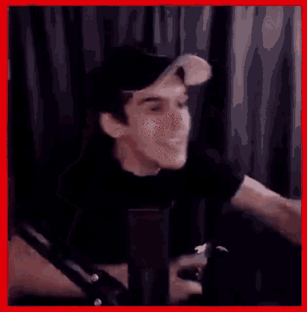 a man wearing a baseball cap is standing in front of a black curtain and making a funny face .