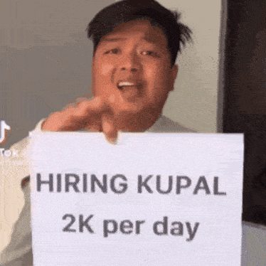 a man is holding a sign that says `` hiring rupal 2k per day '' and pointing at the camera .