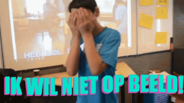 a boy in a blue shirt is covering his face with his hands and the words ik wil niet op beeld behind him