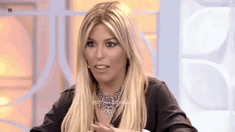 a blonde woman wearing a necklace and a black shirt is sitting in front of a wall .