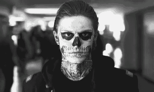 a man with a skull painted on his face and a tattoo on his neck is standing in a hallway .