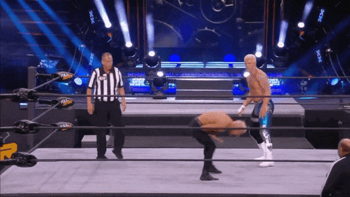 a referee stands behind a wrestler in a wrestling ring while the wrestler performs a trick