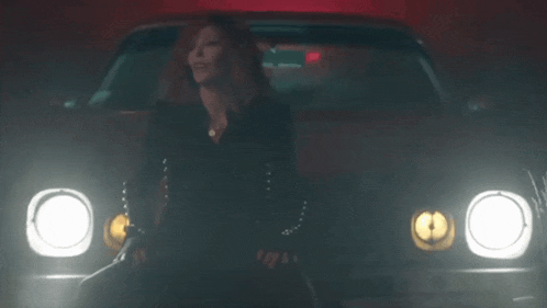a woman is standing in front of a car at night with the headlights on .
