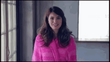 a woman in a pink robe is standing in front of a window smiling .