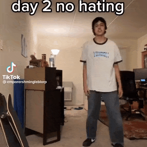 a man in a white shirt is standing in a living room with the words day 2 no hating on the bottom .