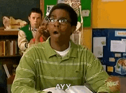 a man in a green shirt and glasses is sitting at a desk in a classroom with his mouth open .
