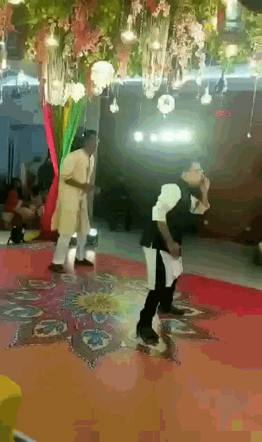 a couple of men are dancing on a red carpet in a room with flowers hanging from the ceiling