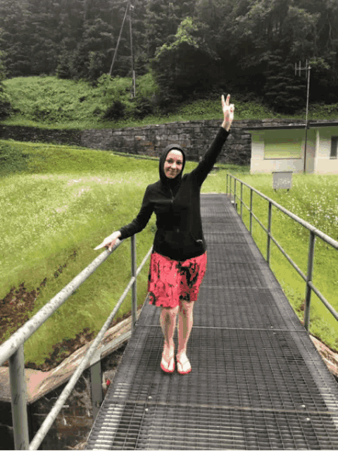 a woman in a black jacket and red shorts stands on a metal bridge giving the peace sign
