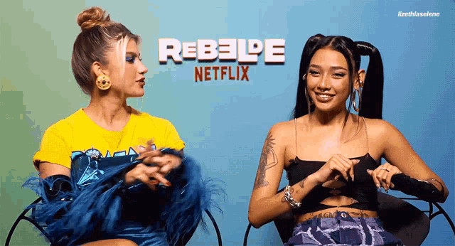 two women are sitting next to each other in front of a rebelde netflix poster