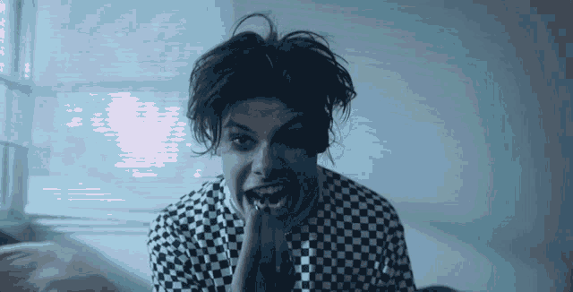 a young man wearing a black and white checkered shirt is sitting on a bed and making a funny face .