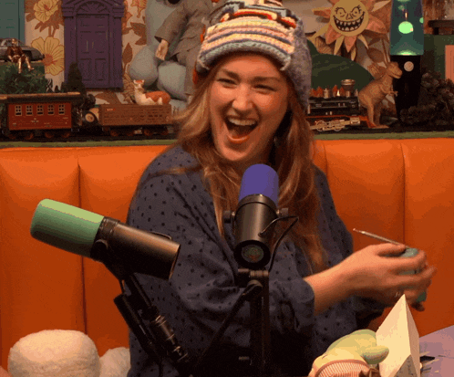 a woman wearing a knitted hat laughs in front of a blue microphone
