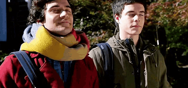 two young men standing next to each other with their eyes closed . one of the men is wearing a scarf around his neck .