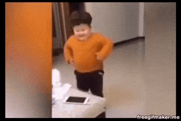a little boy in an orange sweater is standing in a room with a tablet on the floor .