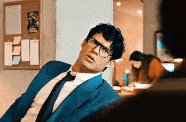 a man in a blue suit and tie is sitting in front of a bulletin board with papers on it .