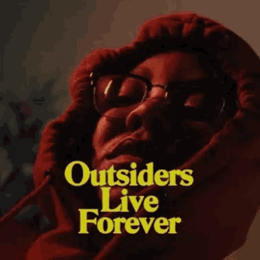 a person wearing glasses and a red hoodie with the words `` outsiders live forever '' on it .