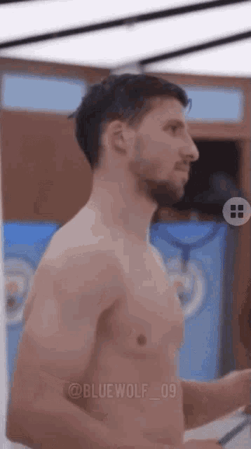 a shirtless man with a beard is standing in front of a mirror in a locker room .