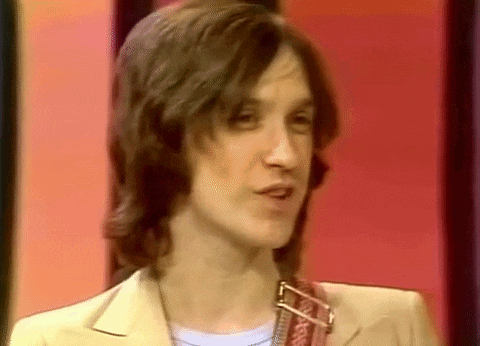 a man with long hair is standing in front of a red wall and looking at the camera .