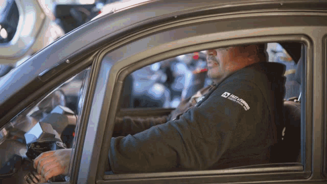 a man wearing a black shirt that says race armored vehicles is sitting in the driver 's seat of a car