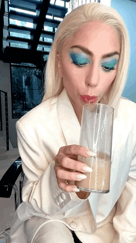 a woman with blonde hair and blue eye shadow is drinking from a tall glass