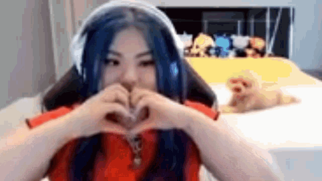 a woman with blue hair is making a heart shape with her hands while sitting on a bed .