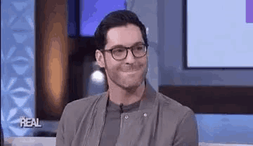 a man wearing glasses is smiling while sitting on a stage in front of a television .