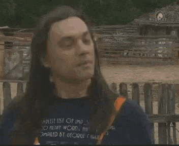 a man with long hair is standing in front of a wooden fence and holding his hair .