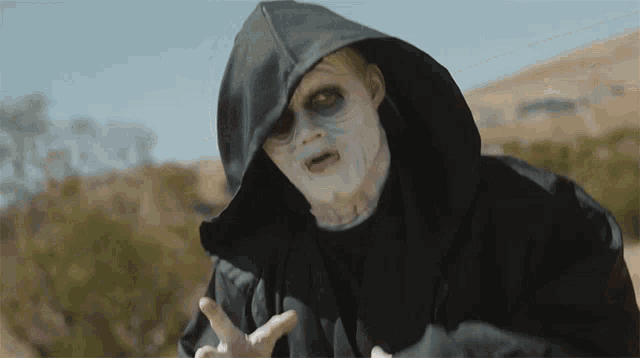 a man in a black robe with a hood and white face paint is standing in the desert .