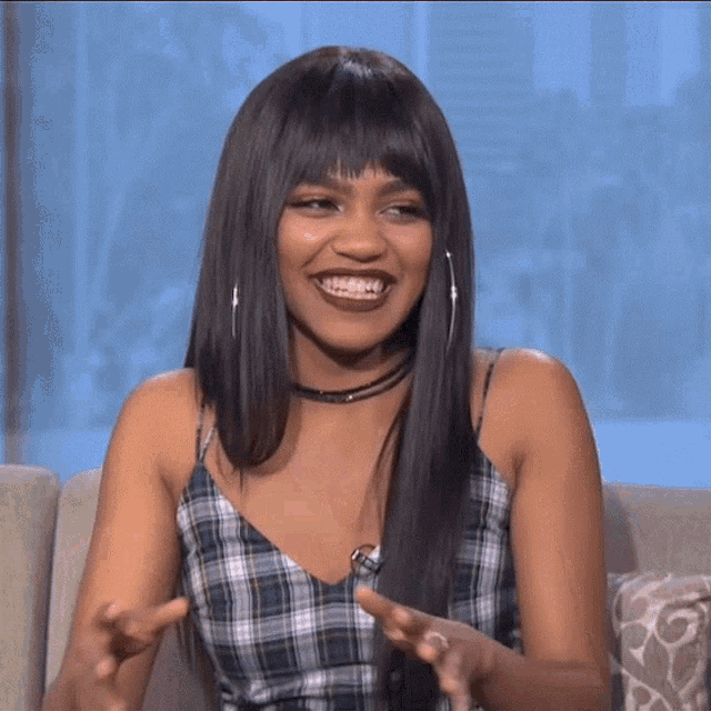 a woman wearing a plaid top and hoop earrings laughs while sitting on a couch
