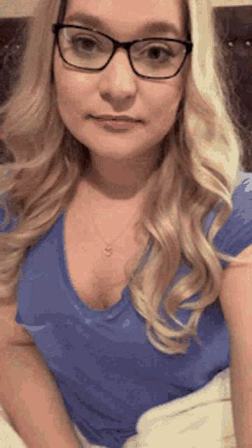 a blonde woman wearing glasses and a blue shirt is sitting on a bed