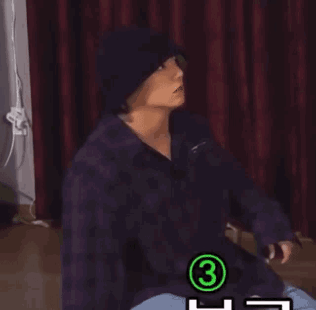 a person wearing a purple plaid shirt and a black hat is sitting on the floor .