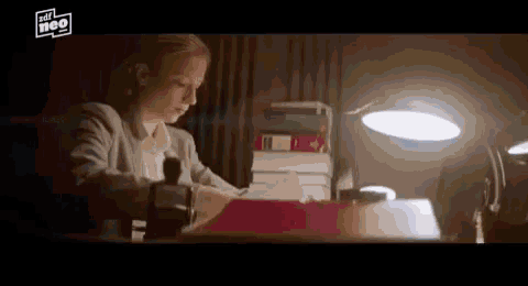 a woman is sitting at a desk with a lamp on and a stack of books .