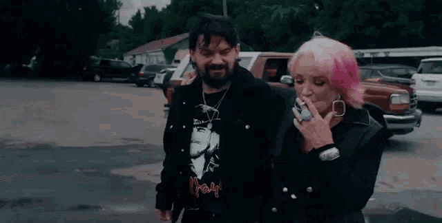 a man and a woman are standing next to each other in a parking lot smoking cigarettes .