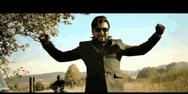a man in a suit and sunglasses is standing in a field with his arms outstretched and a motorcycle in the background .