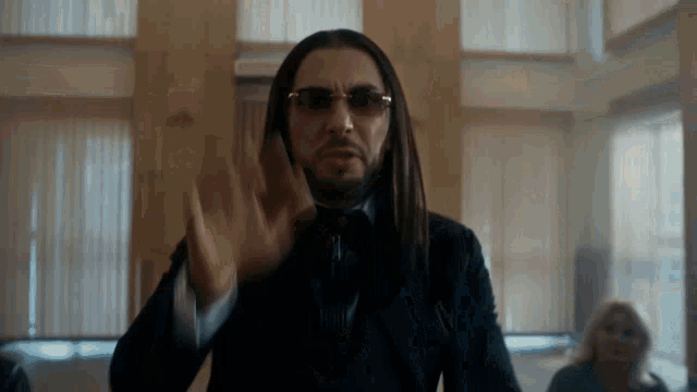 a man with long hair wearing sunglasses and a suit is waving his hand