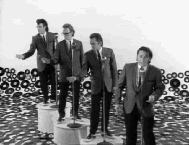 four men in suits and ties are singing into microphones in front of a wall of records