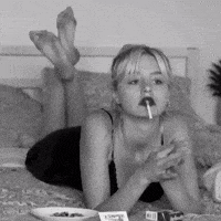 a woman is laying on a bed smoking a cigarette and eating a lollipop .