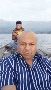 a man in a striped shirt is taking a selfie with another man in a boat in the water .