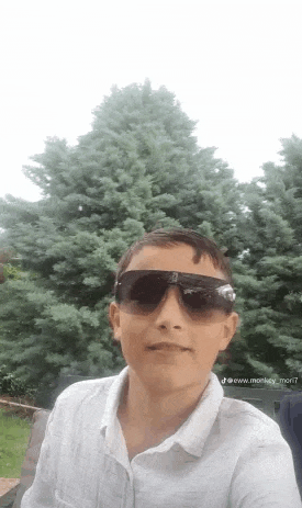 a young boy wearing sunglasses and a white shirt is taking a selfie in front of a tree .
