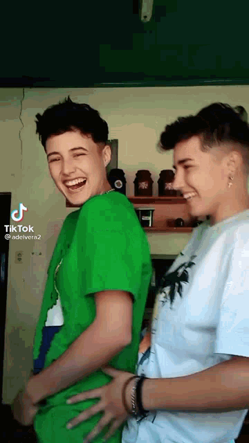 two young men are standing next to each other in a room and laughing . one of the men is wearing a green shirt .