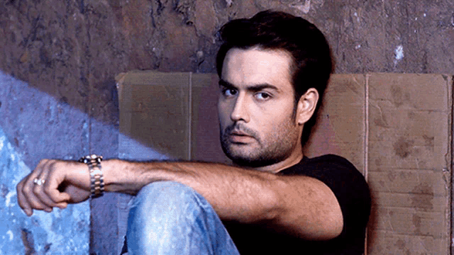 a man wearing a watch sits on a cardboard box against a wall