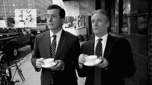 two men in suits and ties are walking down a sidewalk drinking coffee .