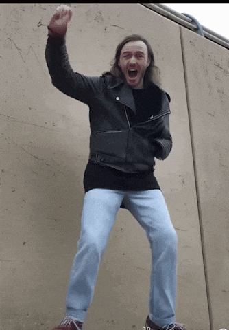 a man wearing a black leather jacket and blue jeans is standing in front of a wall