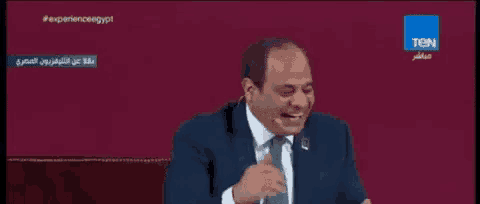 a man in a suit and tie is laughing while sitting in front of a screen that says tcn