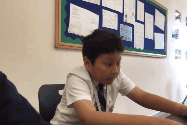 a boy sits at a desk in front of a bulletin board with papers on it