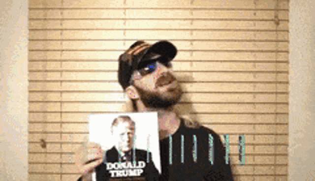 a man wearing sunglasses and a hat holds up a picture of donald trump