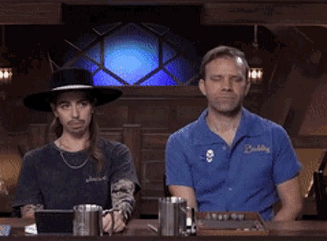 two men sitting at a table with one wearing a blue shirt that says buddie