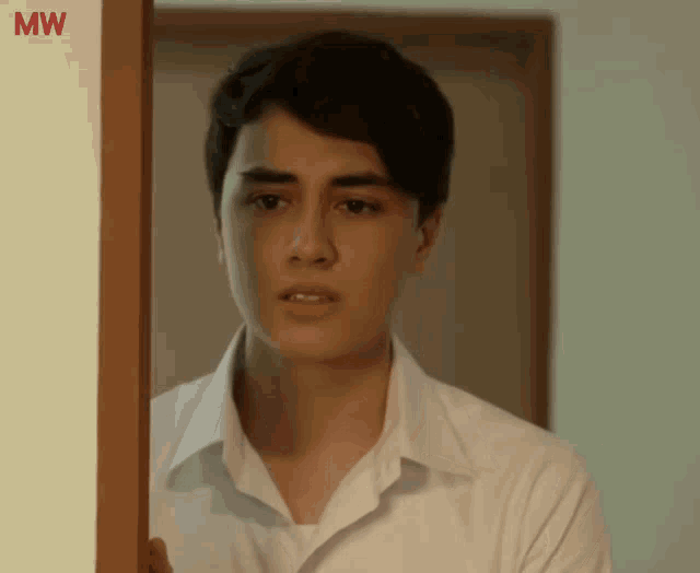 a young man in a white shirt stands in front of a door with mw written on the wall